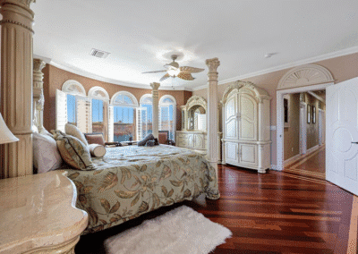 Spacious bedroom with ornate furniture, a large bed, arched windows, and polished wood floors. Sunlight streams in through the windows, and an open door leads to a hallway.