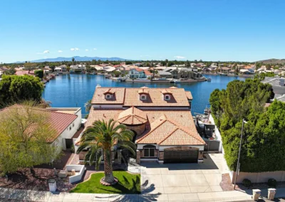 Aerial view of a suburban neighborhood with tile-roofed houses by a lake, palm trees, and distant mountains under a clear blue sky.