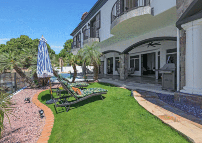 Lounge chairs and an umbrella are set on a grassy lawn beside a modern house with a covered patio, grill, and pool in the background.