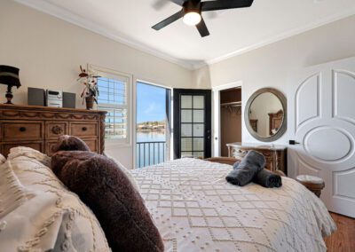 A bedroom with a bed, dresser, and mirror, featuring light walls, ceiling fan, and a door opening to a balcony with a water view.