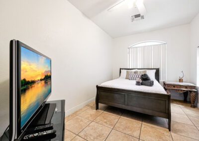 A bedroom with a double bed, bedside table, TV on a stand, tiled floor, and a window with vertical blinds letting in natural light.