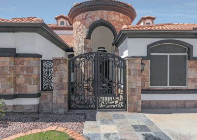 A stone and stucco house features a black wrought iron gate, arched entryway, tiled roof, and large windows.