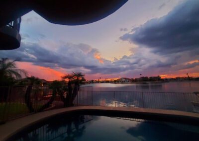 A backyard pool overlooks a calm lake at sunset, with cloudy skies and houses visible across the water.