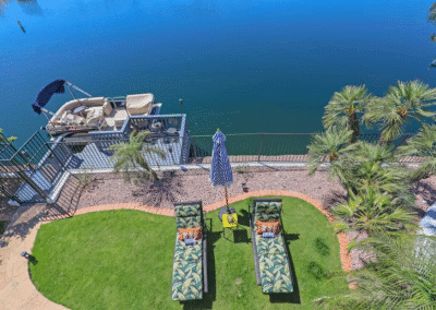 Aerial view of two lounge chairs with cushions on a green lawn near a lakeside docked boat, surrounded by palm trees and a closed umbrella.