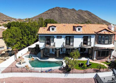A large two-story house with a tile roof, balconies, and a backyard featuring a swimming pool, patio, and mountain views in the background.