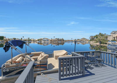 A docked pontoon boat sits beside a gray wooden deck overlooking calm water with houses and palm trees in the background under a clear blue sky.