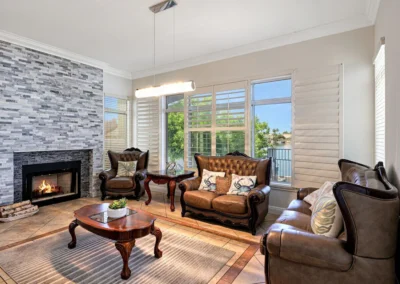 A bright living room with leather sofas, a stone fireplace, a coffee table, large windows with shutters, and a contemporary ceiling light.